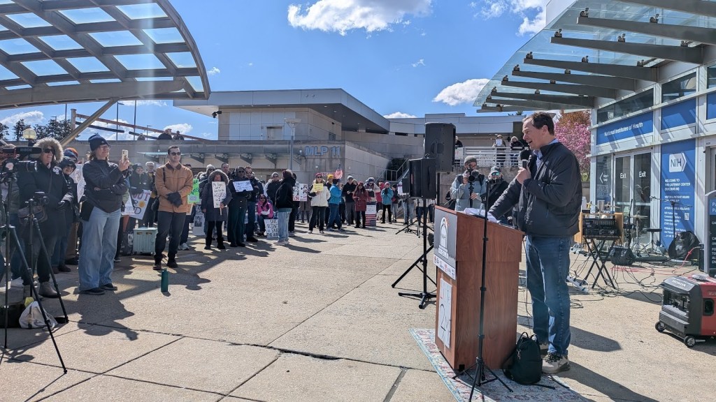 A white man in a navy jacket speaks at a podium in front of NIH.