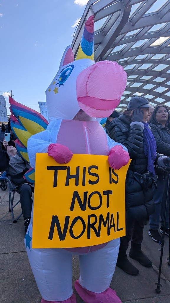 Someone in an inflatable unicorn costume holds a yellow sign with black letters reading "This is not normal"