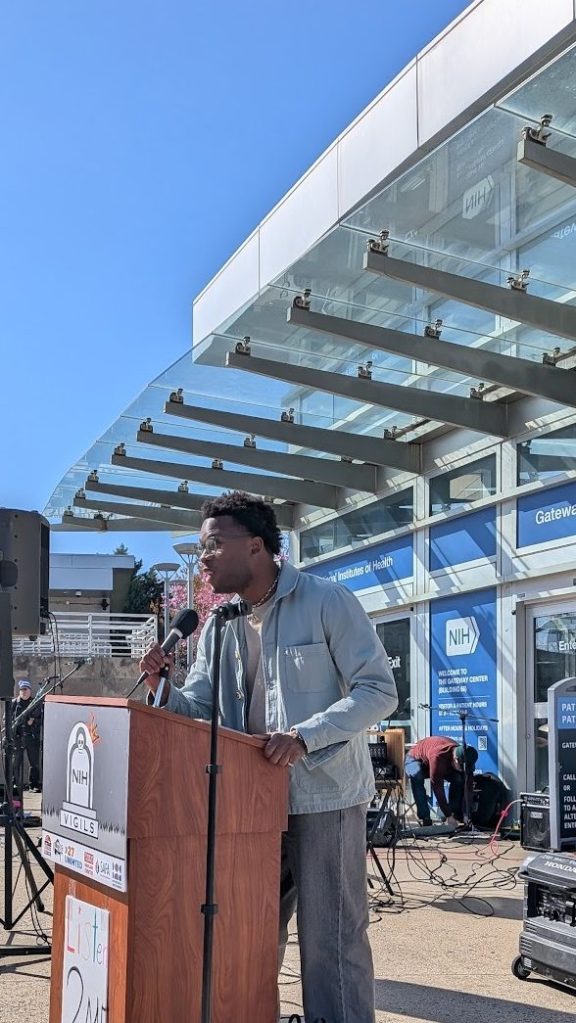 A young Black man in a light-colored denim jacket speaks at a podium in front of NIH.