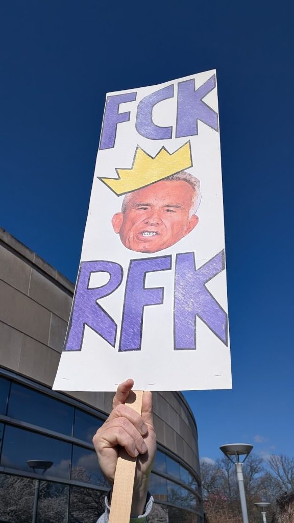 A white sign with blue letters reads "FCK RFK," with a photo of Robert F. Kennedy Jr.'s face, wearing a crown